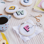 Embroidery hoops with letters 'A', 'D', 'C', 'W', and 'F' on a table with coffee and snacks.