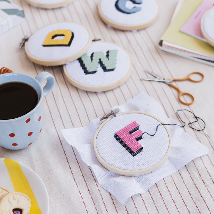 Embroidery hoops with letters 'A', 'D', 'C', 'W', and 'F' on a table with coffee and snacks.