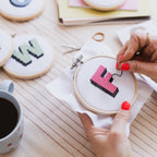 Person embroiderying a small pink and black design on a wooden table with cookies and a mug in the background.