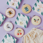 A styled flat-lay of a cross-stitch kit set on a lavender background, featuring embroidered finished pieces in hoop, multiple pattern boxes, and a cream drawstring bag.