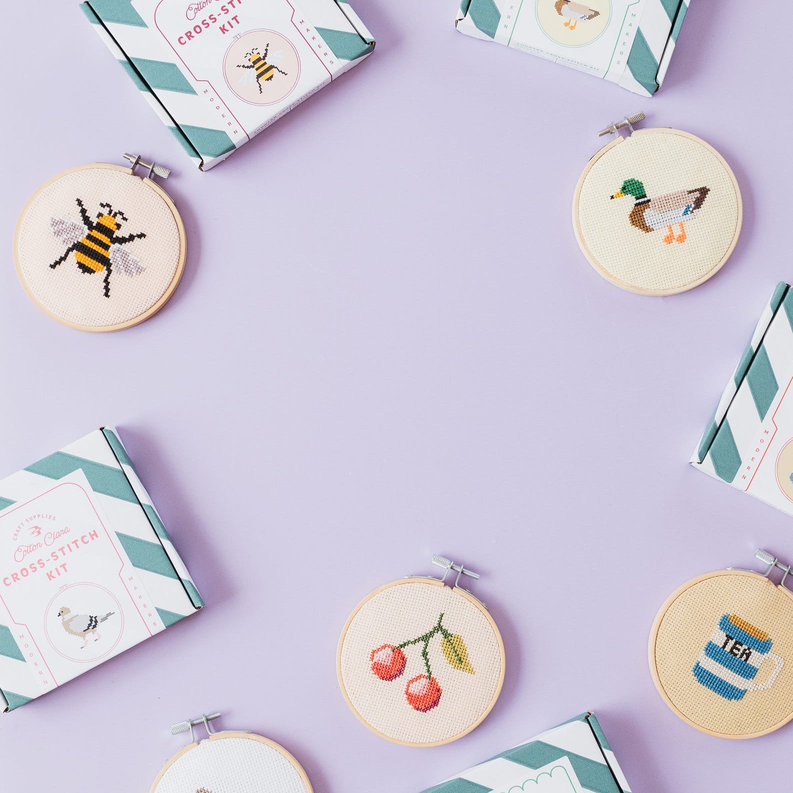 A styled flat-lay of cross-stitch kits and finished embroidered hoops arranged on a light lavender background. The kits and boxes feature mint green and white striped packaging with cream labels, while the round embroidery hoops show completed designs including a bee, duck, cherries, bird, penguin, and striped figure.
