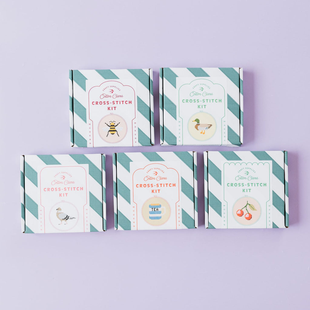 Five 'Cross Stitch Kit' boxes arranged in two rows on a light lavender background. The boxes feature a white and teal diagonal striped border with vintage-style illustrations and text for different cross stitch patterns.