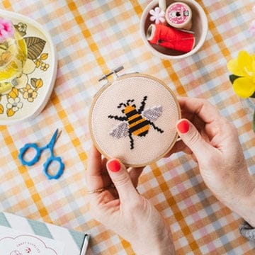 A person's hands holding a circular embroidery hoop with a finished cross-stitch design of a yellow and black bumblebee on a peach fabric background. The scene is set on a yellow, orange, and white checkered tablecloth with a cross-stitch kit book, blue scissors, and other sewing supplies.