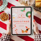 A person holds a tablet displaying a Christmas-themed cross stitch pattern cover titled 'Christmas' by Erin Clima. The scene is styled with a red and white striped tablecloth, a mug of hot chocolate, pretzels, and holiday decorations.