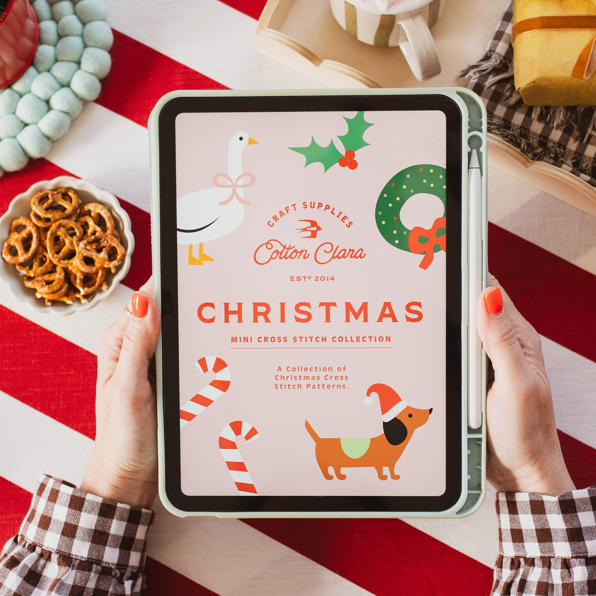 A person holds a tablet displaying a Christmas-themed cross stitch pattern cover titled 'Christmas' by Erin Clima. The scene is styled with a red and white striped tablecloth, a mug of hot chocolate, pretzels, and holiday decorations.