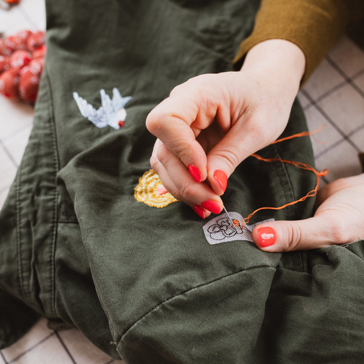 Close-up of a person's hands with red nail polish stitching a small patch onto an olive green garment laid on a white checkered blanket.