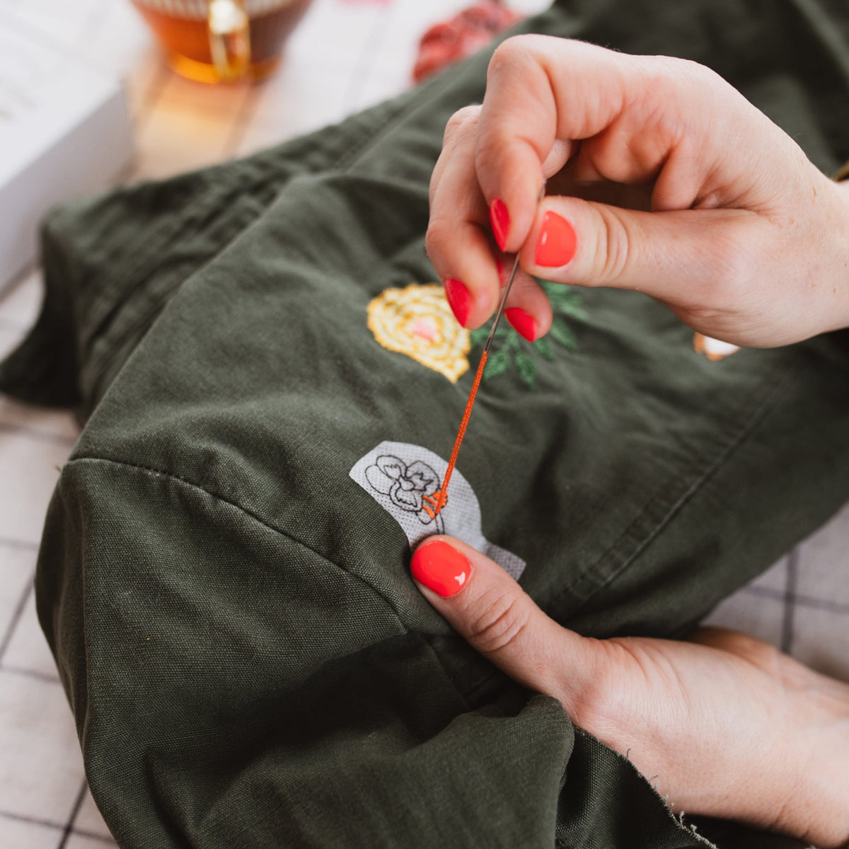 A close-up lifestyle shot of a person's hands with bright coral nail polish stitching an embroidered patch onto an olive green garment. The background is a soft-focus indoor setting with a glass of tea and a checkered floor.