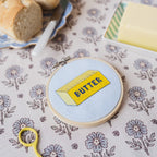 Embroidery hoop with a 'butter' design on a floral tablecloth with bread and coffee.
