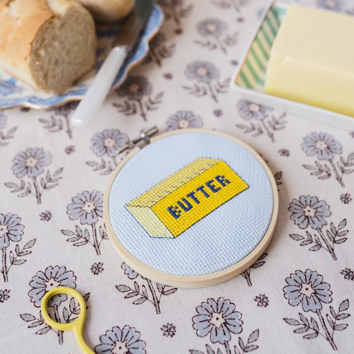 Embroidery hoop with a 'butter' design on a floral tablecloth with bread and coffee.