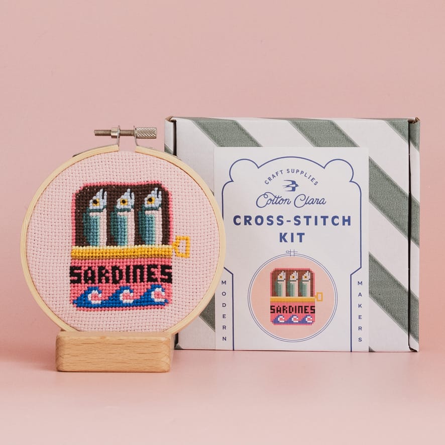 A cotton cross-stitch kit box next to a completed cross-stitch piece showing a sardine boat, displayed on a wooden stand against a solid pink background.