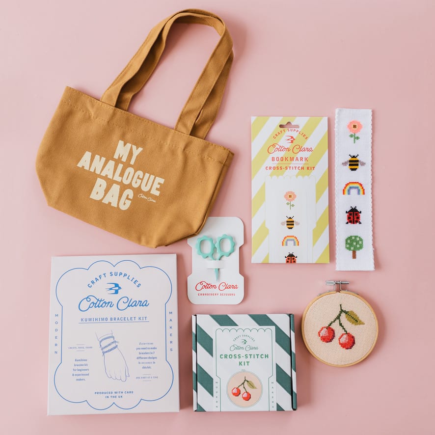 A flat-lay arrangement of a tan canvas tote bag with white text reading 'MY ANALOGUE BAG' alongside a Cotton Clara cross-stitch kit, embroidery scissors, pattern booklets, and an embroidery hoop with cherry stitching, all set against a solid pink background.
