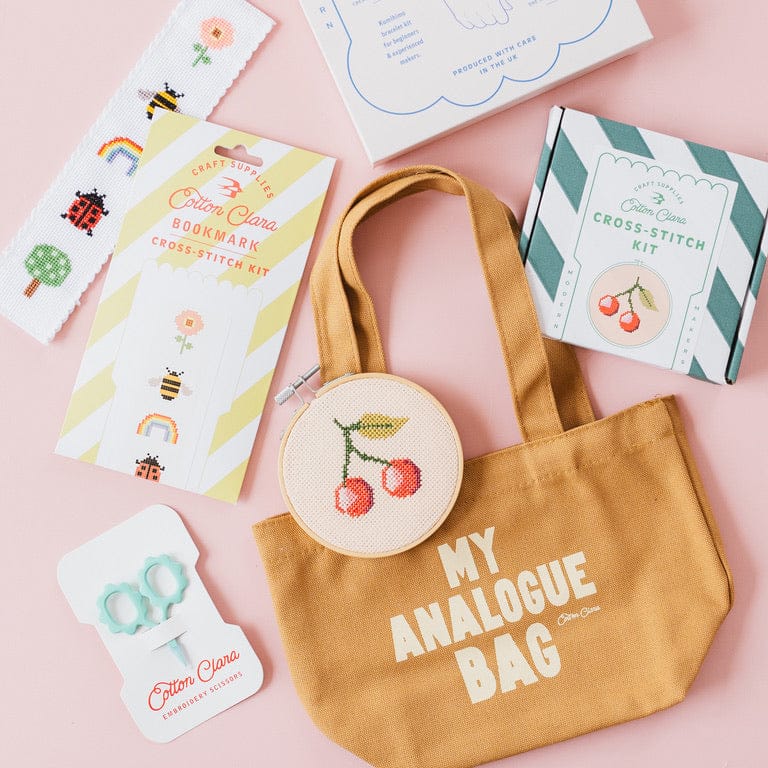 A styled flat-lay of a Cotton Clara 'My Analogue Bag' cross-stitch kit on a solid pink background, including the canvas tote bag, embroidery hoop with cherry design, boxes, and instruction cards.