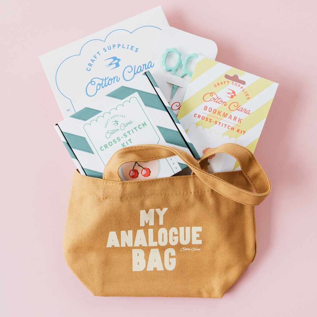 A tan canvas tote bag with the words 'MY ANALOGUE BAG' printed in white on the front, filled with craft supplies including a card that reads 'Cotton Clara' and a booklet, set against a solid light pink background.