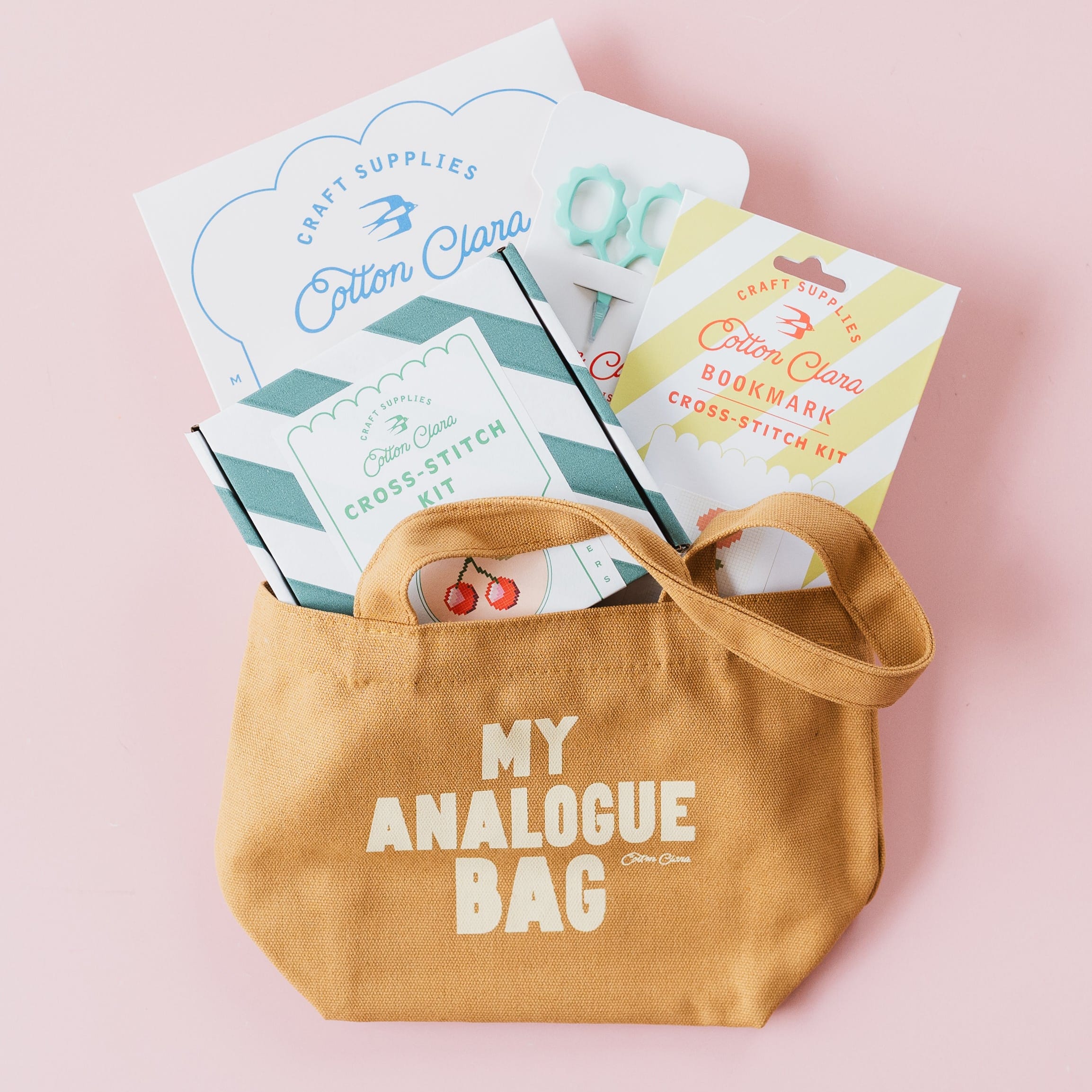 A tan canvas tote bag with the words 'MY ANALOGUE BAG' printed in white on the front, filled with craft supplies including a card that reads 'Cotton Clara' and a booklet, set against a solid light pink background.