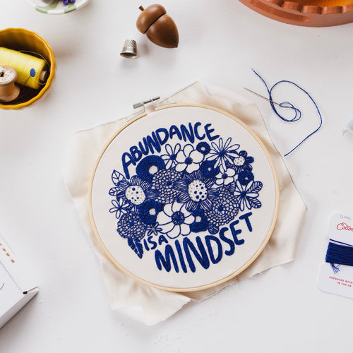 Flat-lay image of stitched Cotton Clara Hoop Club Abundance is a Mindset design, in an embroidery hoop, with a needle and thread still attached, and a thread bobbin by its side